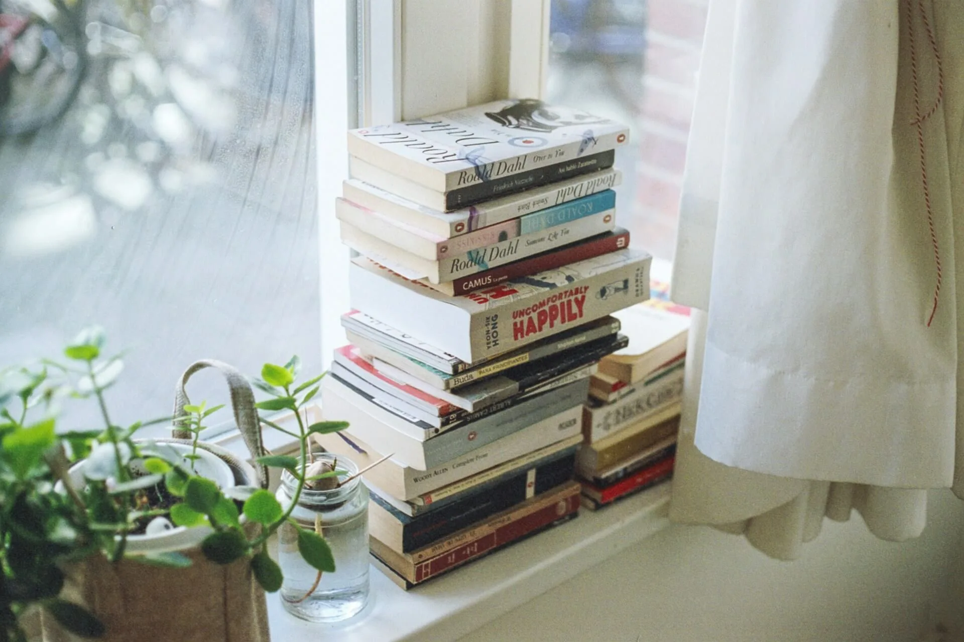 How to read 10 books a month - a stack of well-read books on a windowsill, peaking out from behind a white curtain.